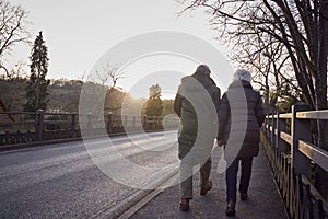 Rear View Of Loving Senior Couple Enjoying Autumn Or Winter Walk Along Road By Bridge