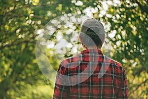 Rear view of male farmer wearing plaid shirt and trucker`s hat standing in walnut orchard and looking at trees