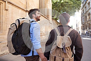 Rear View Of Male Gay Couple On Vacation Wearing Backpacks Holding Hands Walking Along City Street