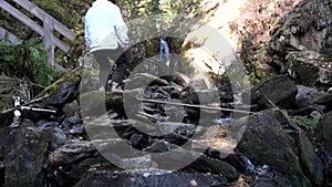 Rear view of a man in hat and white jacket traveling in mountains near small, beautiful waterfall. Footage. Senior man