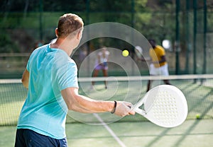 Rear view of man playing paddle tennis match on outdoor court on blurred background of opponents. Sport lifestyle