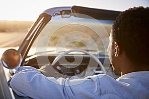 Rear View Of Man On Road Trip Driving Classic Convertible Car Towards Sunset