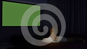 Rear view of a man sitting on sofa in living room and watching tv with green screen. Media. Guy in front of TV with