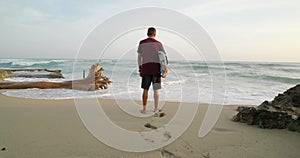 Rear view. Man with surfboard standing on tropical beach against Atlantic Ocean.