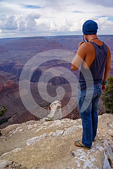 Rear view of man in tank top standing on the edge of a rock on Bright Angel trail with panoramic aerial overlook of South Rim