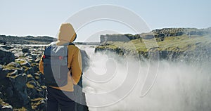 Rear view of man in yellow jacket and backpack standing at cliff edge looking and enjoying at Detifoss waterfall in