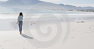 Rear view of mixed race woman walking barefoot on the beach by the sea