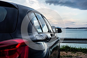 Rear view of modern luxury black car parked above the sea with a romantic view during the sunset
