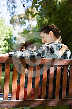 Rear view of mother and daughter sitting with arm around on wooden bench