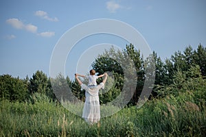 Rear view of mother holding her little son on her shoulders, they walk through field on summer.