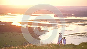 Rear view of mother and little daughter holding hands walks down the hill with perfect view on the river at sunset