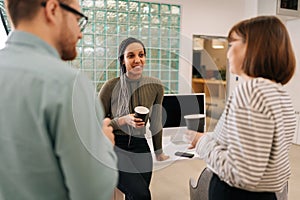 Rear view of multiethnic coworkers standing in office, drinking coffee and talking cheerfully in workspace during break