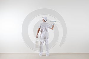 Rear view of painter man looking at blank wall, with paint roller and bucket, isolated on white room