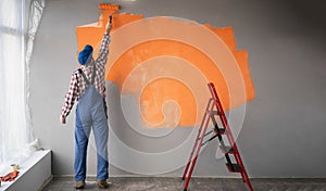 Rear view of painter man painting the wall in orange color with paint roller, empty space with red ladder