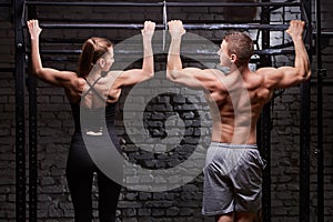 Rear view photo of muscular man and woman doing exercises on horizontal bar against brick wall at the cross fit gym.