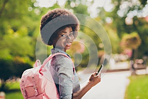 Rear view photo portrait of black skinned female student using smartphone walking in summer park wearing pink backpack