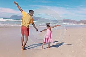 Rear view of playful african american young man and daughter with arms outstretched running at beach