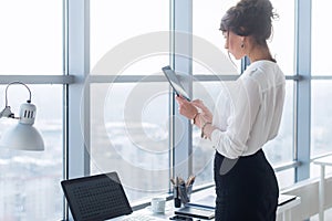 Rear view portrait of a young female office worker using apps at her tablet computer, wearing formal suit, standing near