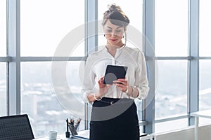 Rear view portrait of a young female office worker using apps at her tablet computer, wearing formal suit, standing near