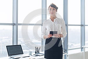 Rear view portrait of a young female office worker using apps at her tablet computer, wearing formal suit, standing near