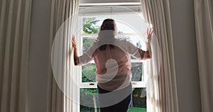 Rear view of senior caucasian woman drawing curtains in sunny room and looking out of window