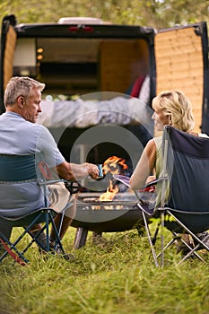 Rear View Of Senior Couple Camping In Countryside With RV Drinking Coffee By Outdoor Fire