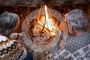 Rear view of senior couple in front of the fireplace while roasting marshmallows. The gray-haired elderly wear winter sweaters