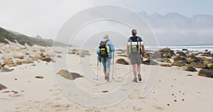 Rear view of senior hiker couple with backpack and hiking poles walking while hiking on the beach