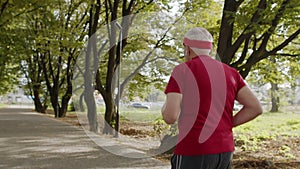 Rear view of senior old man running, working out cardio in park and listening music on earphones