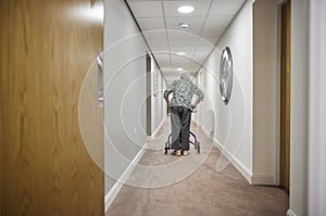 Rear View Of Senior Woman With Walker Walking Along Corridor In Retirement Home