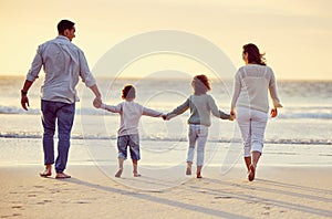Rear view shot of a carefree family holding hands while walking on the beach at sunset. Mixed race parents and their two