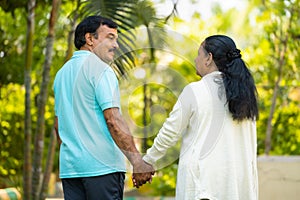 Rear view shot of relaxed senior couple walking at park by holding hands - concept of companionship, healthy actve