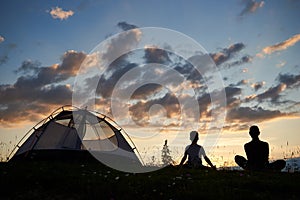 Rear view of silhouette of woman and boy sitting on grass with wild flowers relaxing near the campsite at sunset