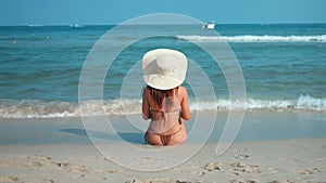 Rear view silhouette of woman in swimsuit sitting on shore in waves of sea and holding hat with hands. Waves lazily wash