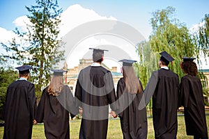 Rear view of six successful international young graduates in black robes and hats finished their education, holding hands and