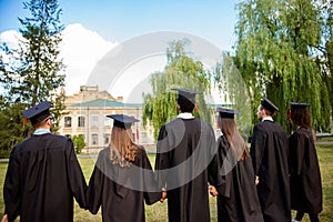 Rear view of six successful international young graduates in black robes and head wear, finished their education, holding hands