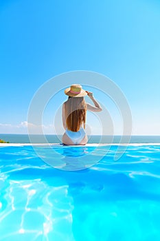rear view. a slender woman in a white swimsuit in a straw hat sits on the pool.