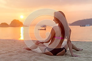 Rear view of slim female model sitting on seashore wearing bikini looking away during sunrise with sun path reflected