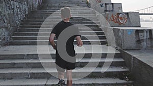 Rear view of slim fit athlete running on quay. Sportive active man runs along river. Athletic young boy running on promenade near