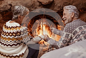 Rear view of smiling senior couple in front of the fireplace while roasting marshmallows hand in hand. The gray-haired elderly