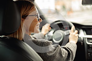 Rear view of smiling woman driving car and holding both hands on steering wheel on the way to work