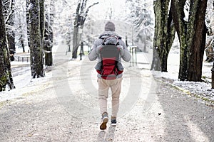 Rear view of sporty father carrying his infant son wearing winter jumpsuit and cap in backpack carrier walking in city