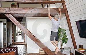 Rear view of a sporty and healthy middle aged man exercising at home, doing pull ups on wooden bar in the living room of