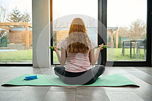 Rear view of sporty pregnant woman with long hair , sitting on lotus position on a yoga mat and exercising with dumbbells looking