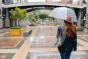 Rear view to unrecognizable redhead young woman in fashion hat walking on European city street with transparent umbrella