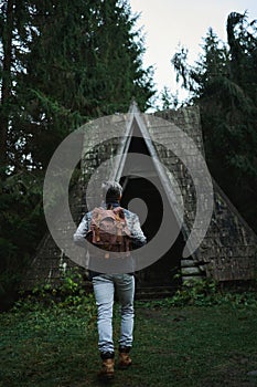 Rear view traveler man with backpack coming to old vintage wooden hut in dark woods