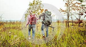 Rear view of a traveling couple of backpacker wading outdoors through an autumn field holding hands with hiking poles