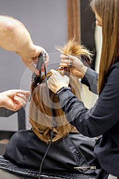The rear view of two hairdressers are curling hair for a young woman with electric hair iron in a beauty salon.