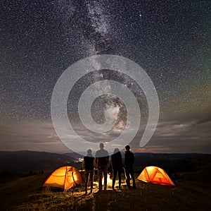 Rear view two young couples at night in tent camp stand enjoying starry sky, Milky way, mountains and luminous town