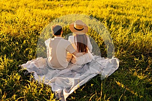 Rear view of unrecognizable young couple in love hugging sitting on beautiful field of green grass in sunset sunlight on